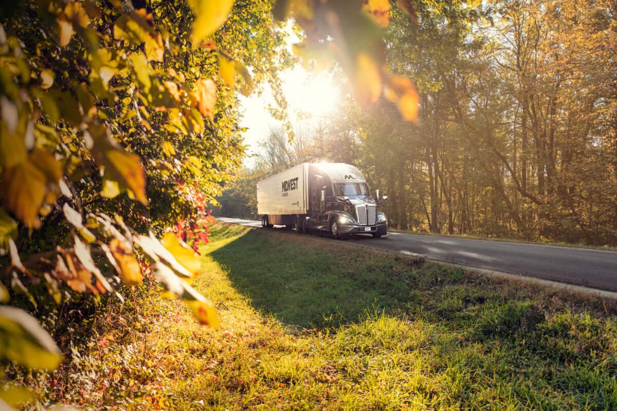 A Midwest Carriers truck on the highway with the sun shining above the truck and fall leaves on the side of the road.