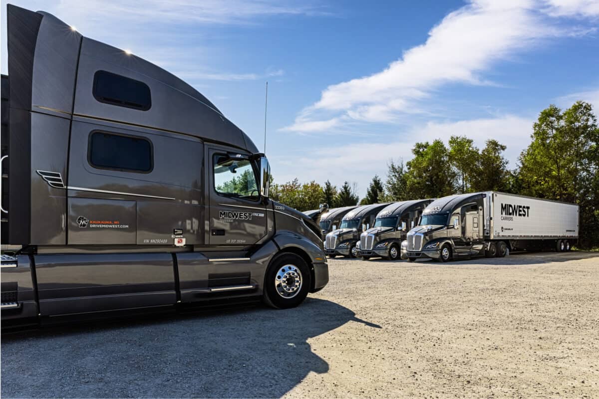 Gravel parking lot with six Midwest Carrier trucks lined up with tree line in the background