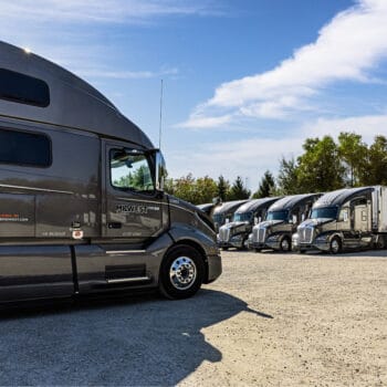 Gravel parking lot with six Midwest Carrier trucks lined up with tree line in the background
