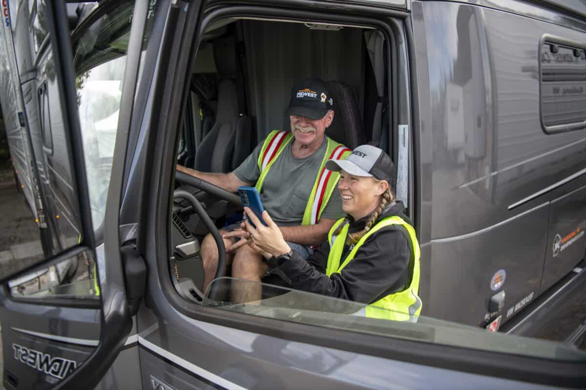 One Midwest Carriers driver sits in behind the wheel of a semi, while another stands beside them, pointing to features within the cab of the truck.
