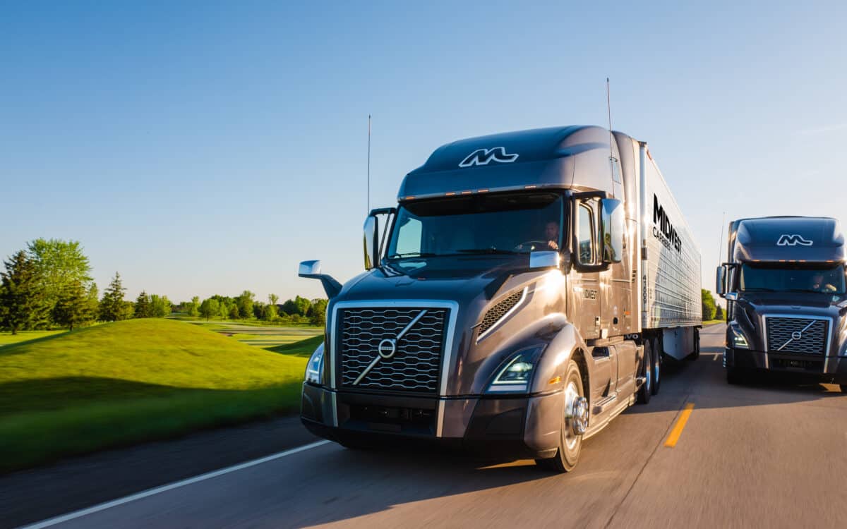 Two Midwest Carriers trucks drive side by side along a road with rolling hills and trees in the background