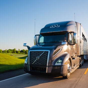 Two Midwest Carriers trucks drive side by side along a road with rolling hills and trees in the background