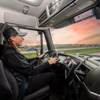 Female truck driver driving a Midwest Carriers truck down a split highway with cars driving by.