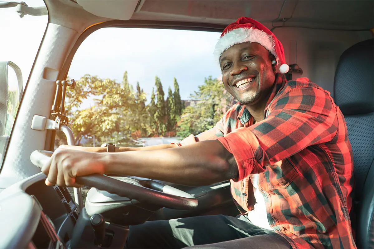 A male truck driver in plaid clothing driving a truck wearing a santa hat.